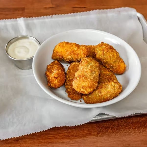 A white bowl of golden-brown fried pickles with a side of ranch dressing on a white paper-lined tray.